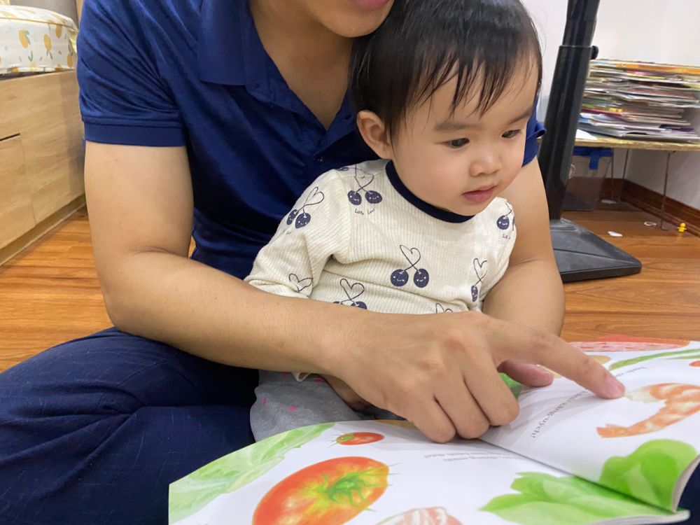 Ba đang đọc sách tranh cho bé 1 tuổi nghe, bé chăm chú lắng nghe và nhìn vào tranh